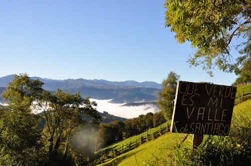 Paisaje de montaña de Asturias, con la niebla cubriendo el fondo del valle. En primer plano se ve un cartel con el texto «Que verde es mi valle C de Santiago»